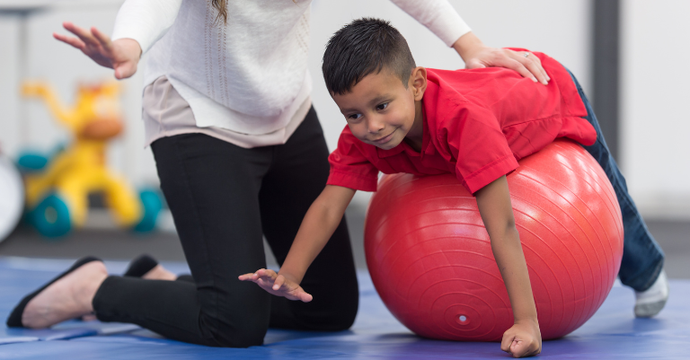 child in red t-shirt doing physical therapy on a red exercise ball, aided by a PT